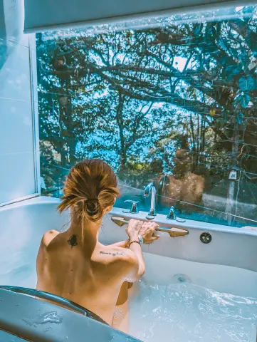 AprilJakovish, seen from behind, sits naked in a bubbling hot tub looking out a large window at green trees.