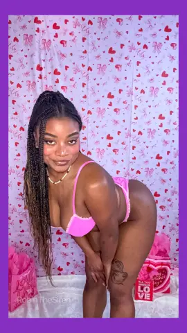 Curvy Black woman in pink lingerie posing playfully against a romantic red heart patterned backdrop.