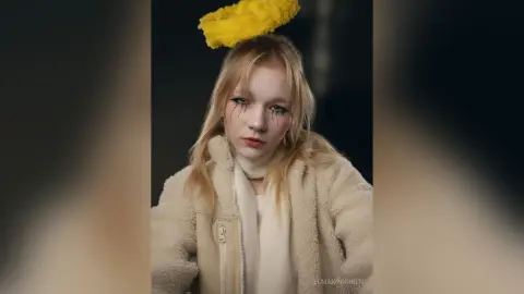Blonde woman in a cozy cream fleece jacket and white scarf posing with a dreamy expression against a dark background.