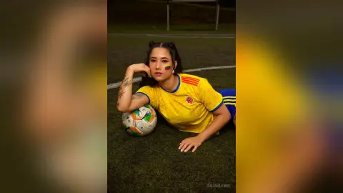 JuliahLewis lies on a grass field wearing a yellow soccer jersey with a soccer ball tucked under her arm.
