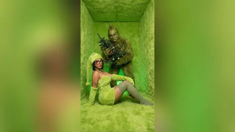 Woman in green fuzzy outfit and hat sitting next to Grinch character in a lime green textured room.