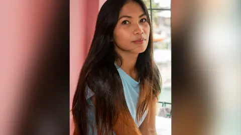 Serene young woman with long dark hair wearing a light blue top in a bright, sunlit room.