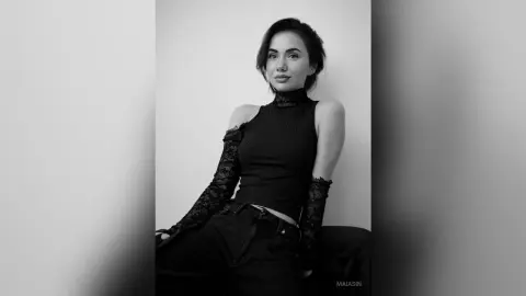 Elegant woman in a black lace mock neck top and dark trousers posing in a minimalist studio setting.