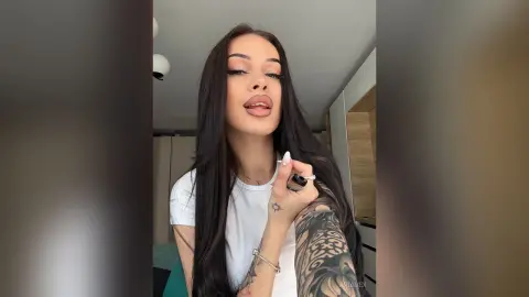 AriaVex poses with long dark hair and a white t-shirt, showing off her large arm tattoo.