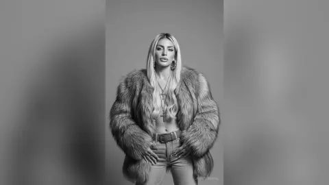 JessycaPage poses in a large fur coat and jeans, looking directly at the camera with long blonde hair.