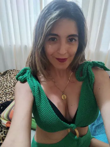 Smiling woman in a green knit crop top and gold necklace relaxing in a bright indoor setting.