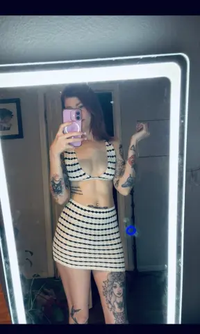 Tattooed woman in a cream striped bikini top and mini skirt posing in a lit mirror for a casual selfie.
