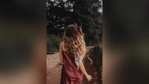 Woman in a red satin slip dress standing by a tranquil lake in a moody forest setting.