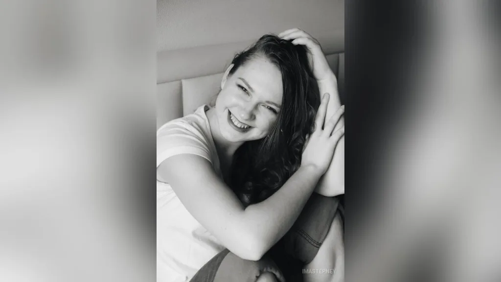 ImaStepney sits on a bed wearing a white t-shirt and denim shorts, looking to the side with her hair down.