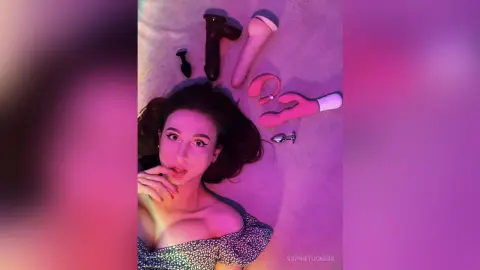 Young woman in floral top lying on bed surrounded by adult toys in moody pink lighting.