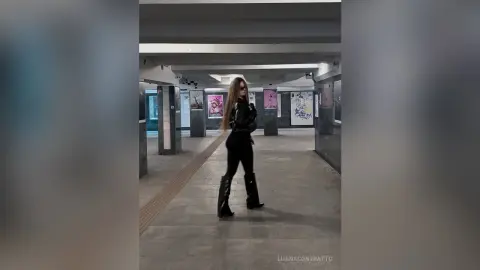 LuanaContratto stands in a hallway wearing a black leather jacket and black leggings with tall black boots, letting her long brown hair hang down.