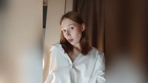 Young woman in a white button-down shirt looking thoughtfully in a soft-lit indoor setting.