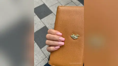 Hand holding a tan pebbled leather designer clutch wallet with gold hardware on a checkered floor.