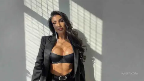 Confident woman in a black blazer and bra top posing against a sunlit wall with dramatic shadows.