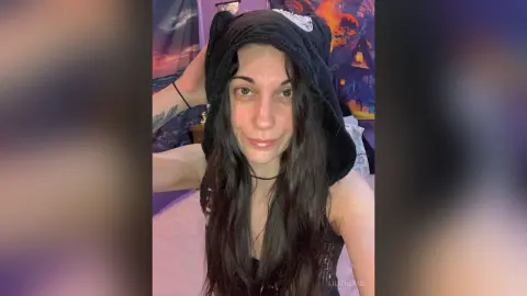 Smiling woman with long dark hair wearing a cozy black hooded animal hat in a dimly lit bedroom.