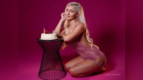 Anita kneels next to a white cake on a black stand, wearing a sheer pink mesh bodysuit that shows her breasts and skin.