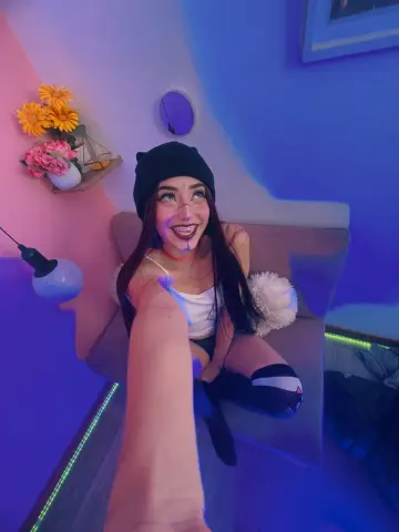 Smiling woman in a white tank top and black beanie sitting on a sofa in moody blue and pink lighting.