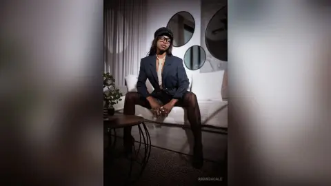 Stylish woman in a navy blazer, black hat, and glasses sitting on a white sofa in a modern living room.