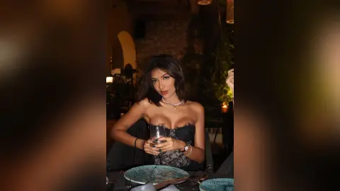 Elegant woman in a black lace dress sitting at a dimly lit restaurant table with a glass of white wine.