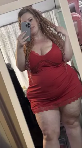 Curvy woman with long wavy hair wearing a red lace chemise nightgown in a bedroom mirror selfie.