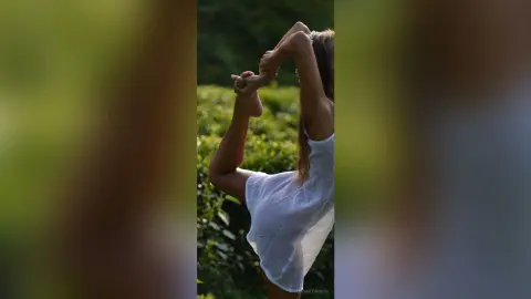 Serene woman in a white summer dress performing a yoga pose in a lush green garden.