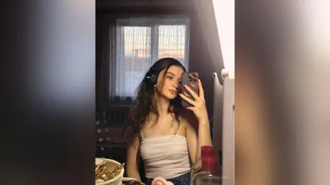 Young woman in a white tank top taking a mirror selfie with headphones in a cozy, sunlit bedroom.