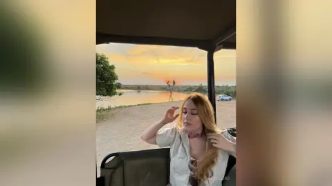 Redheaded woman in a white linen shirt relaxing in a safari vehicle during a peaceful golden hour sunset.