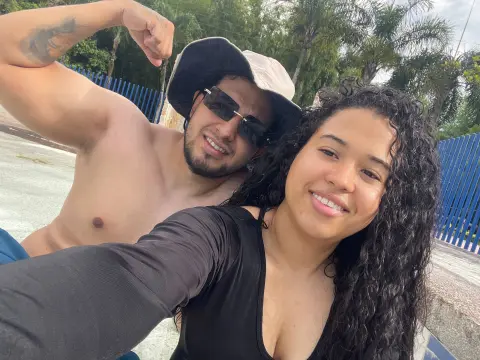 Smiling couple in summer outfits taking a sunny outdoor selfie near palm trees.