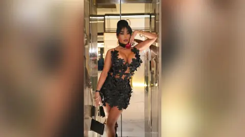 Dolce poses in a black floral lace mini dress with a red rose choker, standing in a bright hallway.