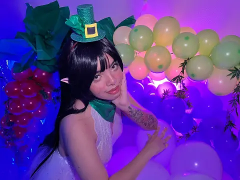 maaeve_elf — Cosplayer in a fairy outfit and leprechaun hat smiling amidst colorful balloons in dreamy purple lighting.