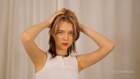 Young woman with blonde hair wearing a white ribbed tank top posing against a neutral curtain background.