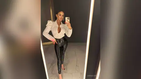 AnnaKarev posing in a mirror wearing a white blouse and tight black shiny pants.