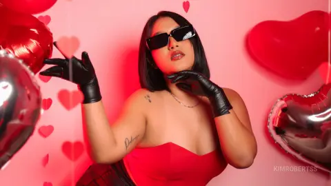 KimRoberth poses in a red strapless top and black gloves, wearing sunglasses against a pink background with red heart balloons.