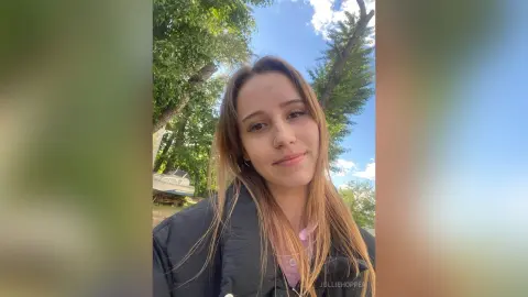 Smiling young woman in a black puffer jacket outdoors under green trees and a blue sky.