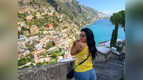 MissyJolie wears a yellow patterned tank top and denim shorts while standing on a stone balcony overlooking a coastal town and blue ocean.