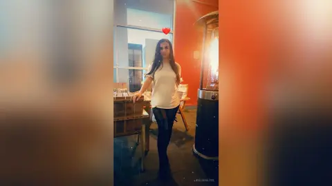 Young woman in a white t-shirt and black jeans standing in a modern restaurant with warm ambient lighting.