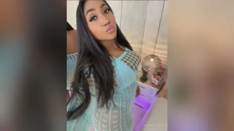 NaiomiLanne poses in a light blue crochet top that shows her skin through the mesh, or open-knit pattern, with her long dark hair down.