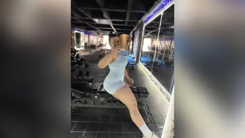 Athletic woman in a white bodysuit taking a mirror selfie in a modern, moody gym setting.