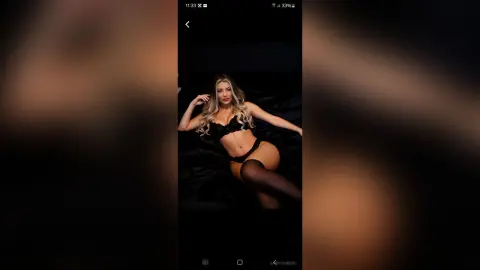 Blonde woman in black lingerie and stockings posing on a dark bed in a moody, intimate setting.
