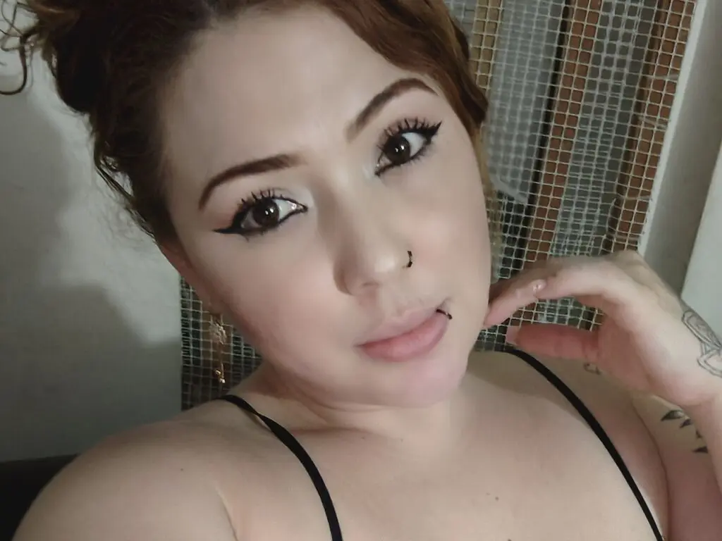 Close-up portrait of a woman with winged eyeliner and nose ring wearing a black camisole in a bright indoor setting.
