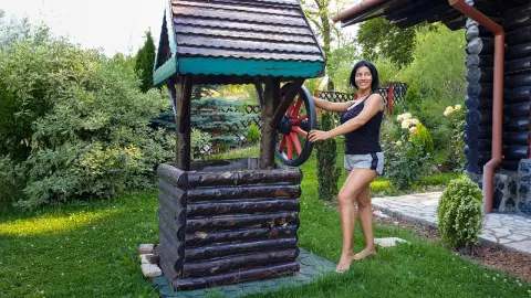 MissyJolie stands next to a wooden wishing well in a garden, wearing a black tank top and grey shorts.
