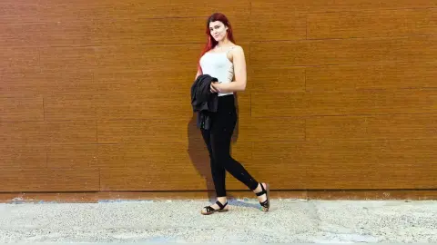Red-haired woman in a white tank top and black leggings posing against a wooden wall in a casual mood.