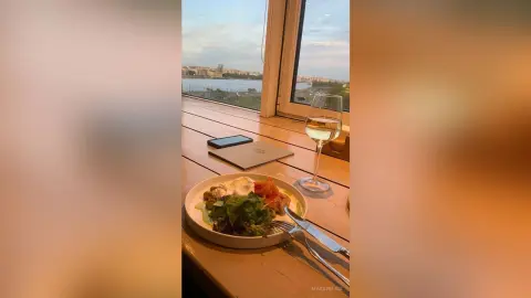 Delicious gourmet salad and white wine on a wooden table overlooking a scenic city river view at sunset.