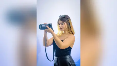 Young woman in a black strapless top holding a professional DSLR camera against a plain blue background.