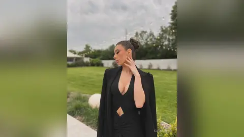 Elegant woman in a black cutout evening gown posing in a lush green garden under an overcast sky.