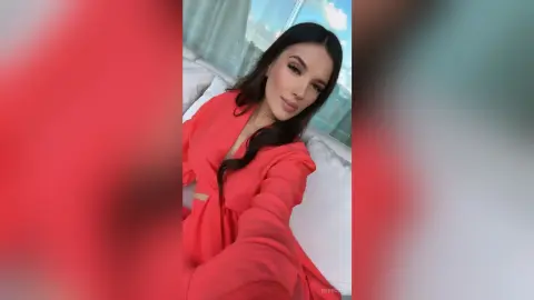 Rebecca poses in a red robe while lounging on a white bed with her long dark hair flowing down.