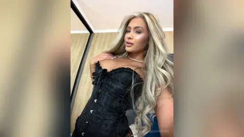 Glamorous blonde woman in a black lace corset dress posing in a modern interior with a confident mood.