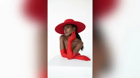 Elegant Black woman in a red wide-brimmed hat and matching opera gloves posing against a white background.