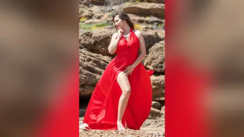 NataliaNacher poses outdoors in a long red dress with a high slit, showing her bare leg while standing against a rocky background.