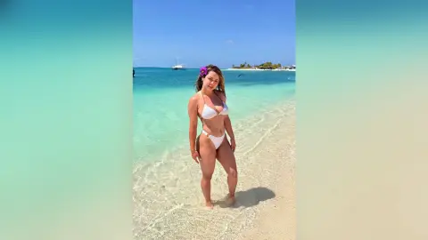 Smiling woman in a white bikini standing on a sunny tropical beach with turquoise ocean water.